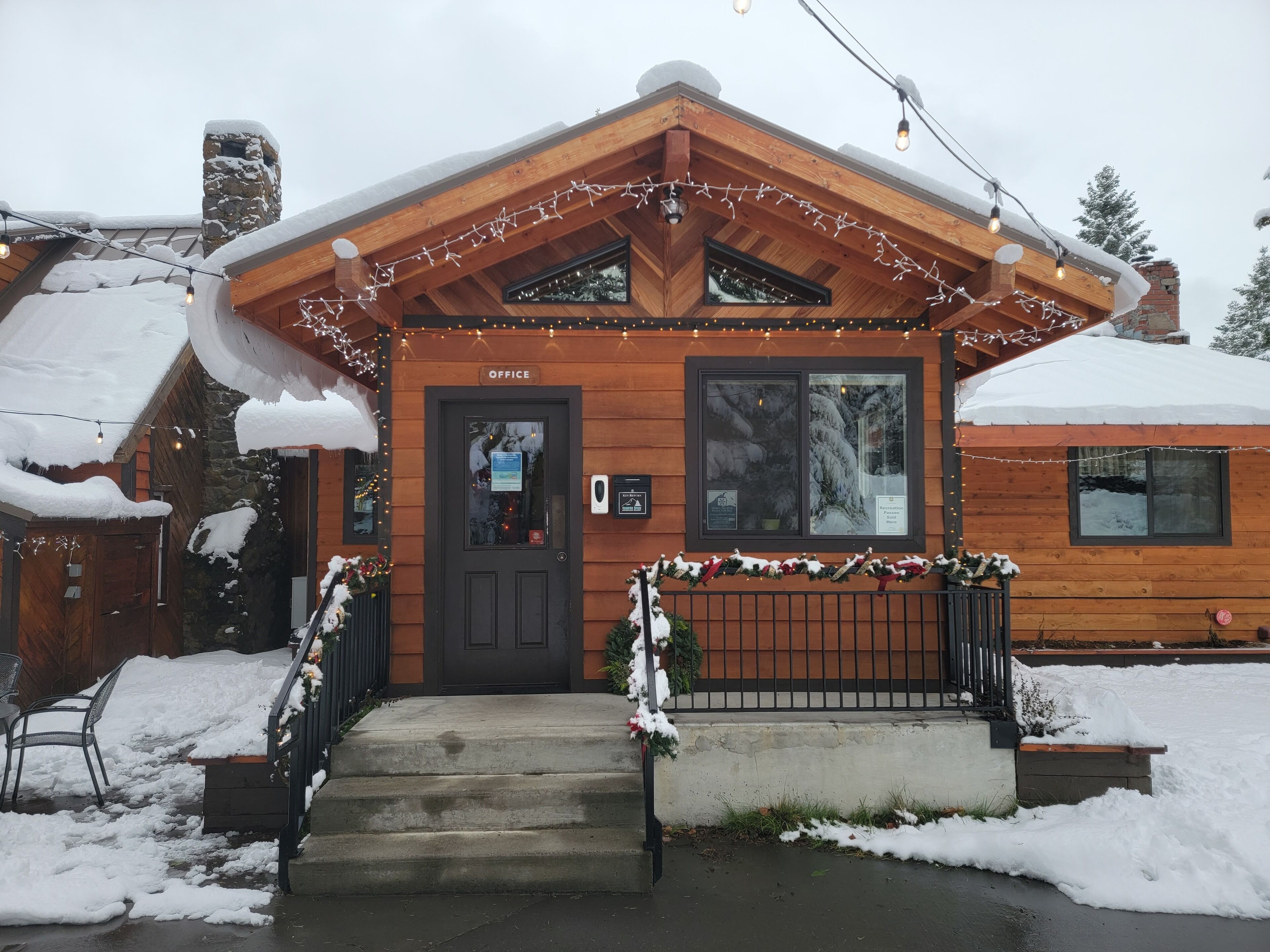 A wooden lodge with a dark entrance door labeled "Office," a stone chimney, and a snow-covered roof.