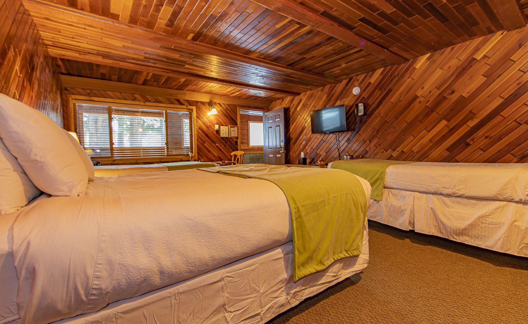 A cozy, wooden-floored room with two beds, a television, and a window with blinds.