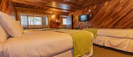 A cozy, wooden-floored room with two beds, a television, and a window with blinds.