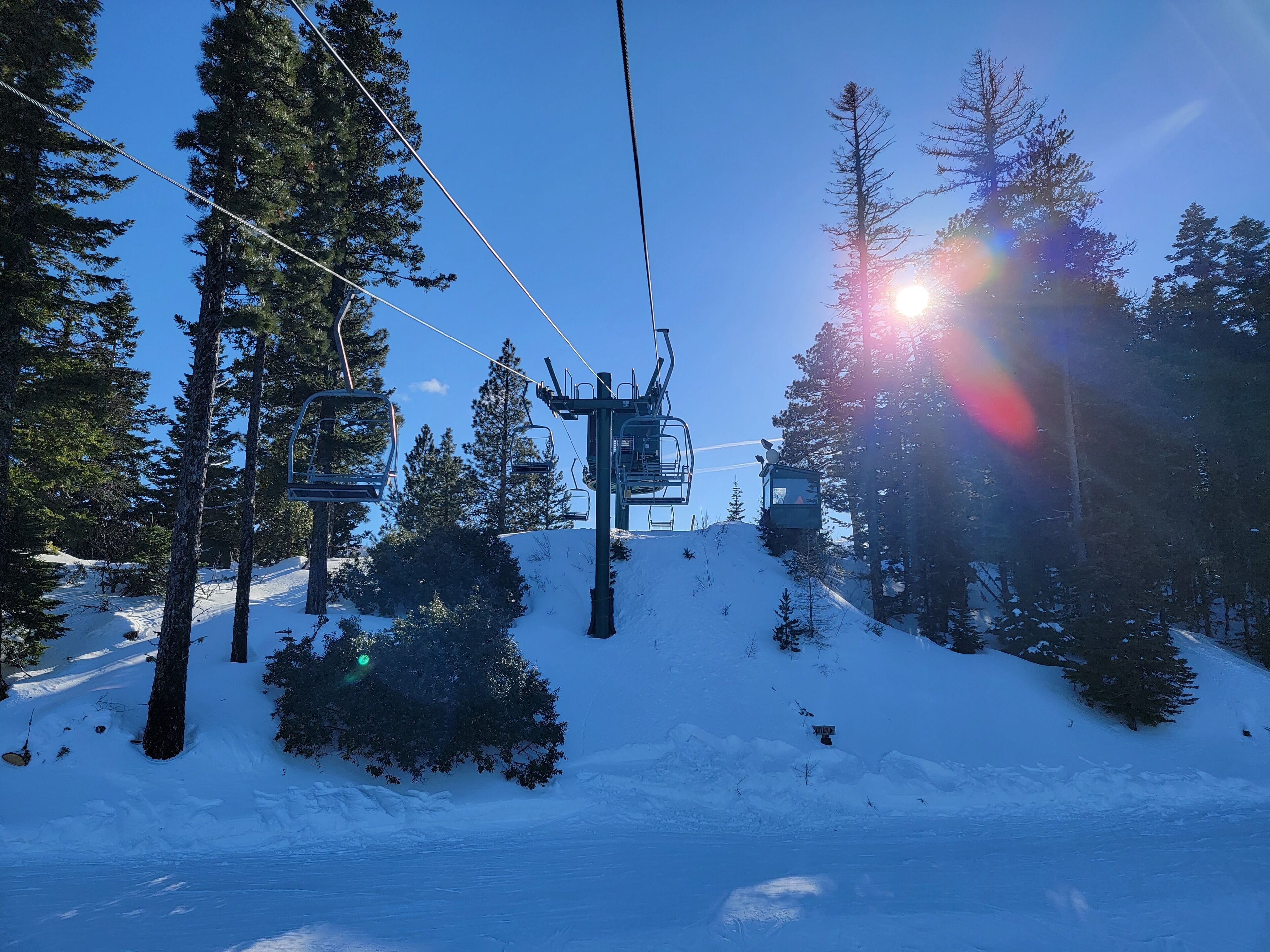 Un télésiège avec des sièges qui montent une pente enneigée, entouré de grands arbres et sous un ciel bleu dégagé.