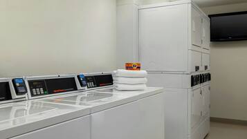 A commercial laundry area with multiple washing machines and dryers, a stack of folded towels, and a wall-mounted television.
