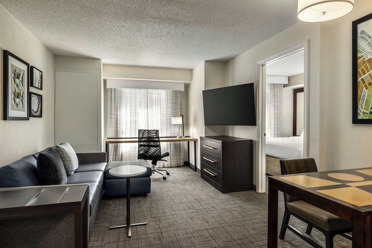 A hotel room with a sofa, a desk, a chair, a TV, and a dining table.