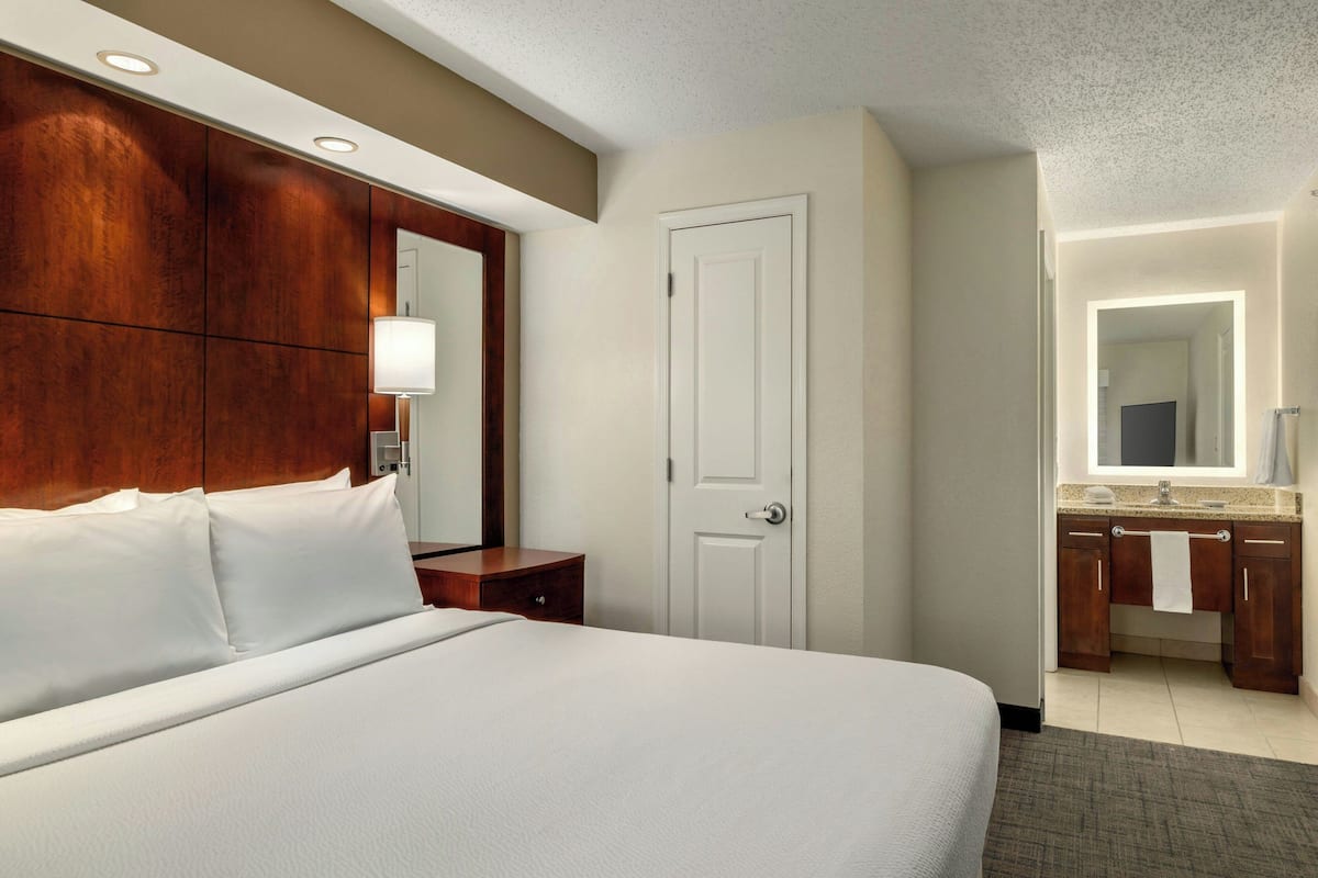 A hotel room with a large bed, a wooden headboard, a nightstand, a bathroom with a sink and mirror, and a door.
