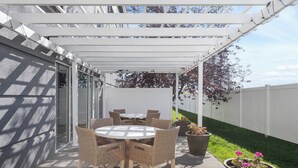 A covered patio area with a round table and wicker chairs, potted plants, and a white fence in the background.