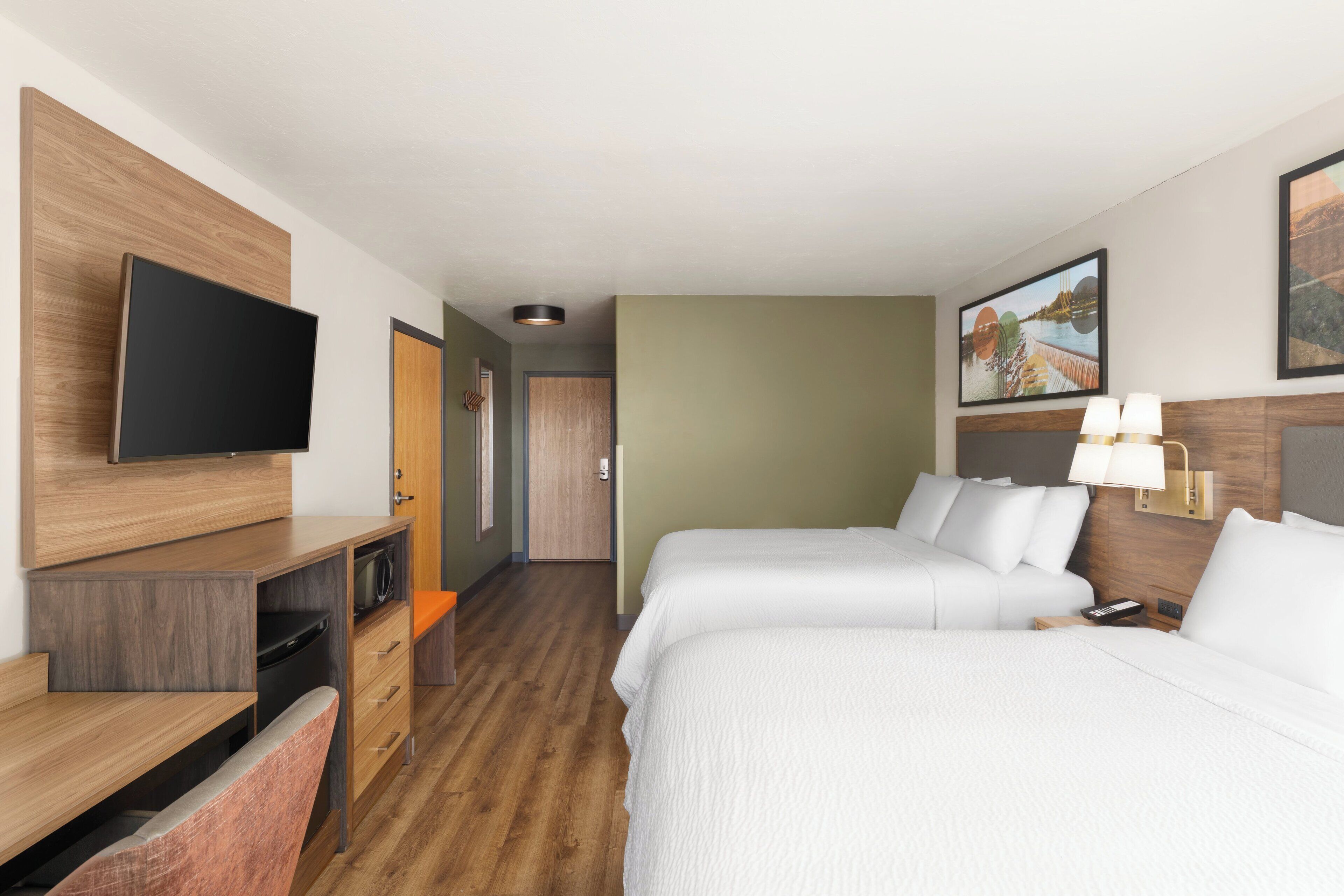 Camera d'albergo con una scrivania in legno, una TV a schermo piatto, due letti, un comodino con una lampada e un quadro appeso al muro.