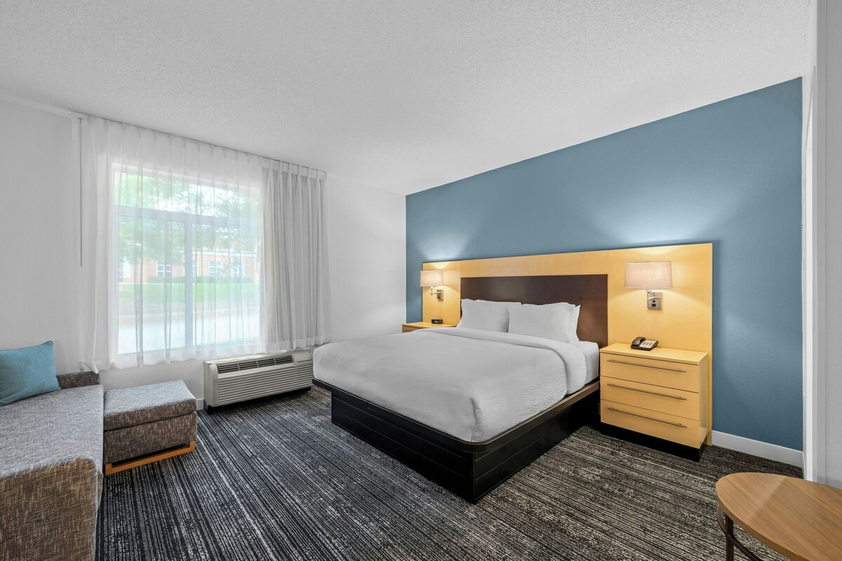 Ein Hotelzimmer mit einem Bett, einem Schreibtisch, einem Sessel, einem Fenster mit Vorhängen und einer Klimaanlage.