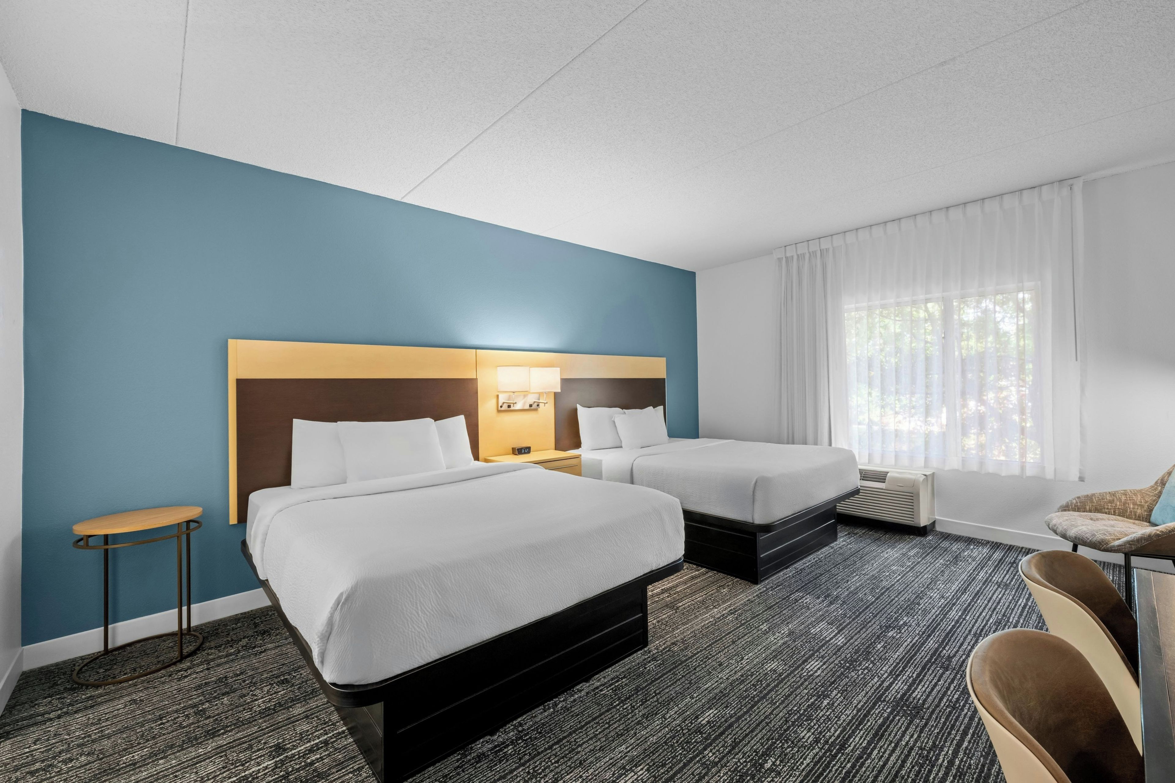 Habitación de hotel con dos camas, una mesita, una silla y un ventanal grande con cortinas.