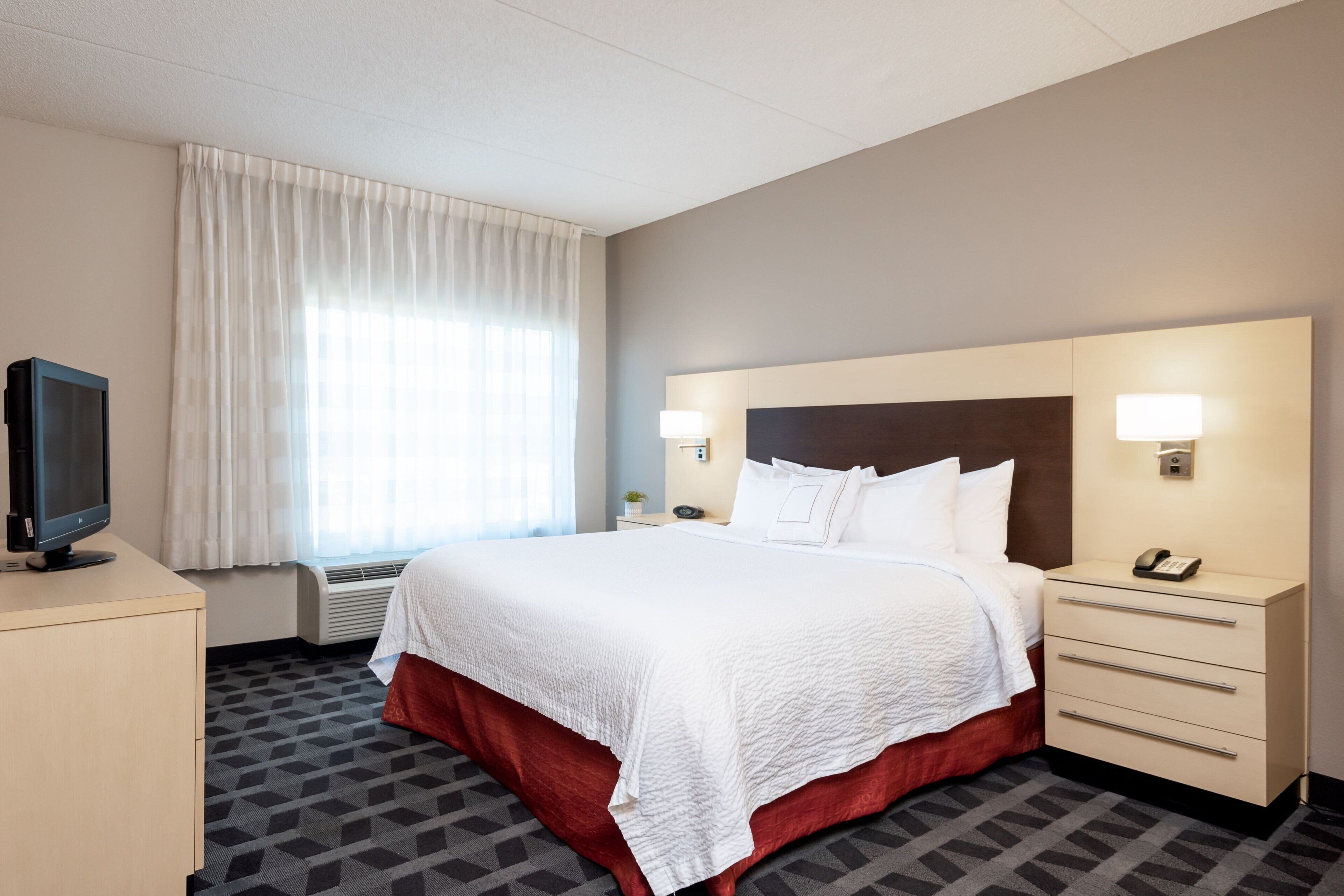 Una habitación de hotel con una cama grande, un escritorio con televisor, una mesita de noche con lámpara y una ventana con cortinas.