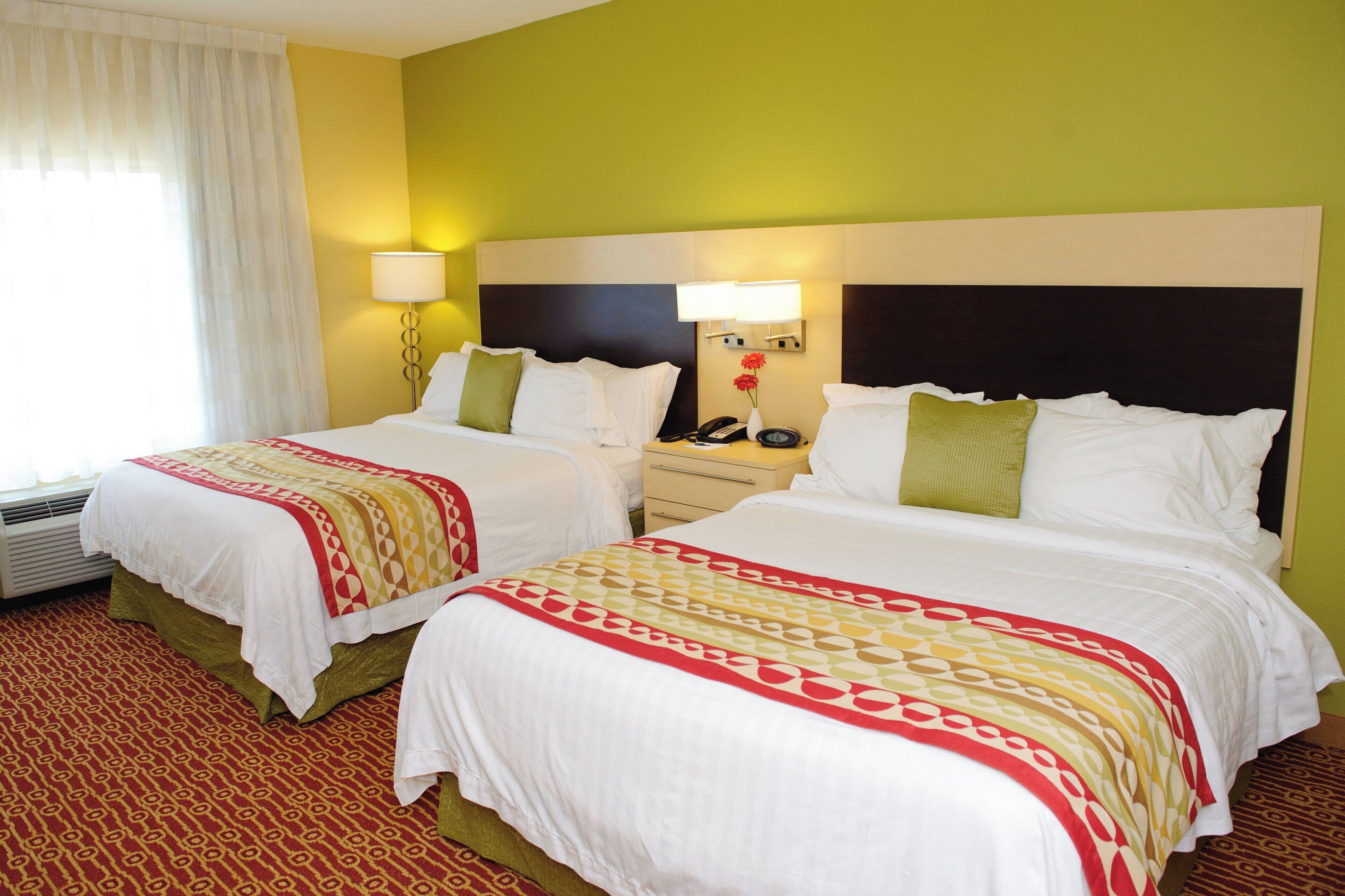 A hotel room with two beds, a nightstand with a lamp, and a window with curtains.