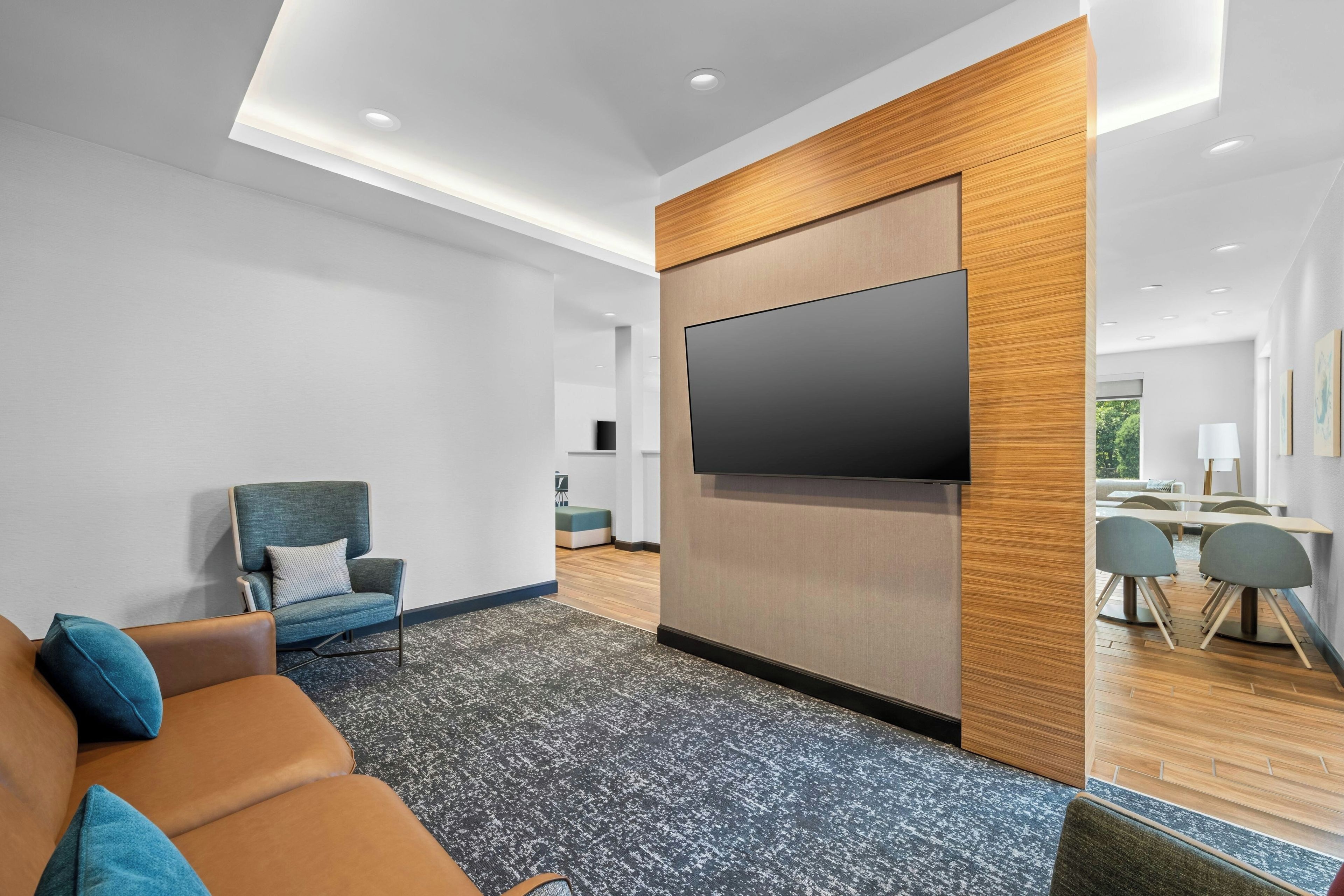 A modern living room with a flat-screen TV mounted on a wooden panel, a comfortable sofa, and a small seating area with a table and chairs.