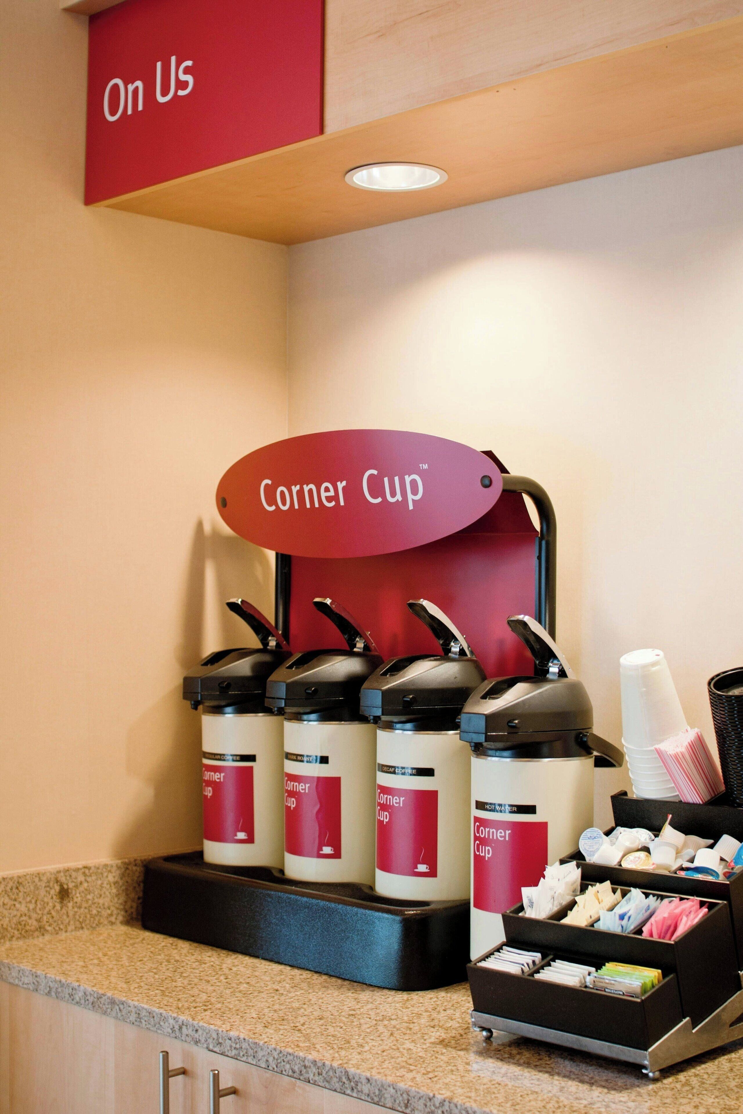 Una estación de café con varios dispensadores de café etiquetados como “Corner Cup” y un letrero que dice “Ofrecido por nosotros”. 