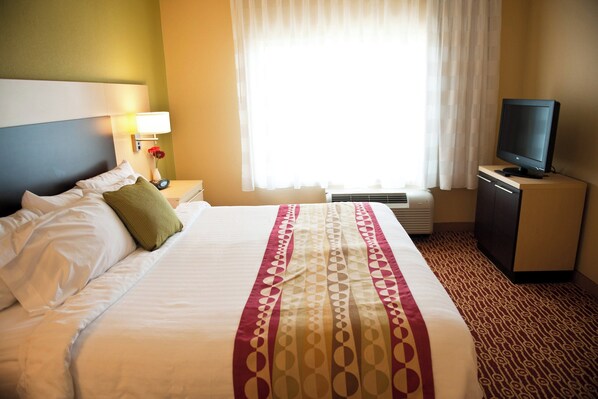 A hotel room with a bed, a television on a stand, a nightstand with a lamp, and a window with curtains.