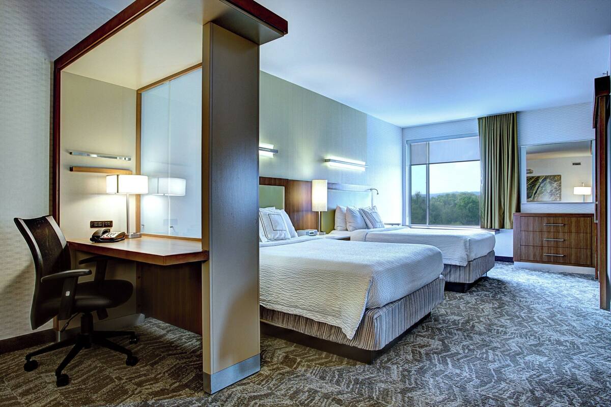 Ein Hotelzimmer mit zwei Betten, einem Schreibtisch mit Stuhl, einem Spiegel und einem Fenster mit Vorhängen.