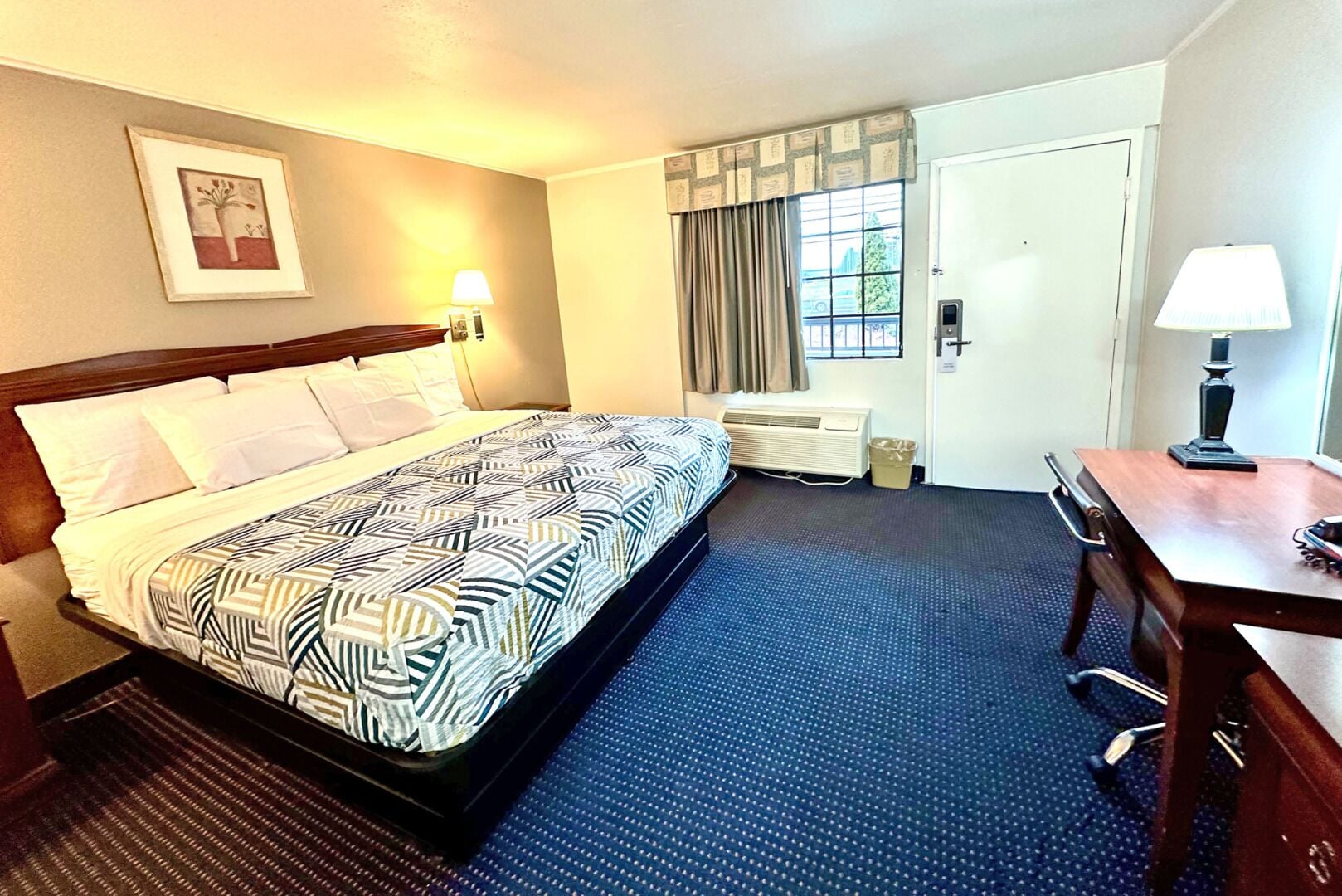 A hotel room with a bed, a desk, a chair, a lamp, a window with curtains, an air conditioning unit, and a framed picture on the wall.