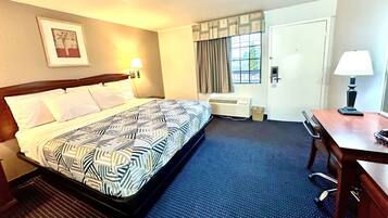 Quarto de hotel com cama, escrivaninha, cadeira, luminária, janela com cortinas, aparelho de ar condicionado e quadro na parede.