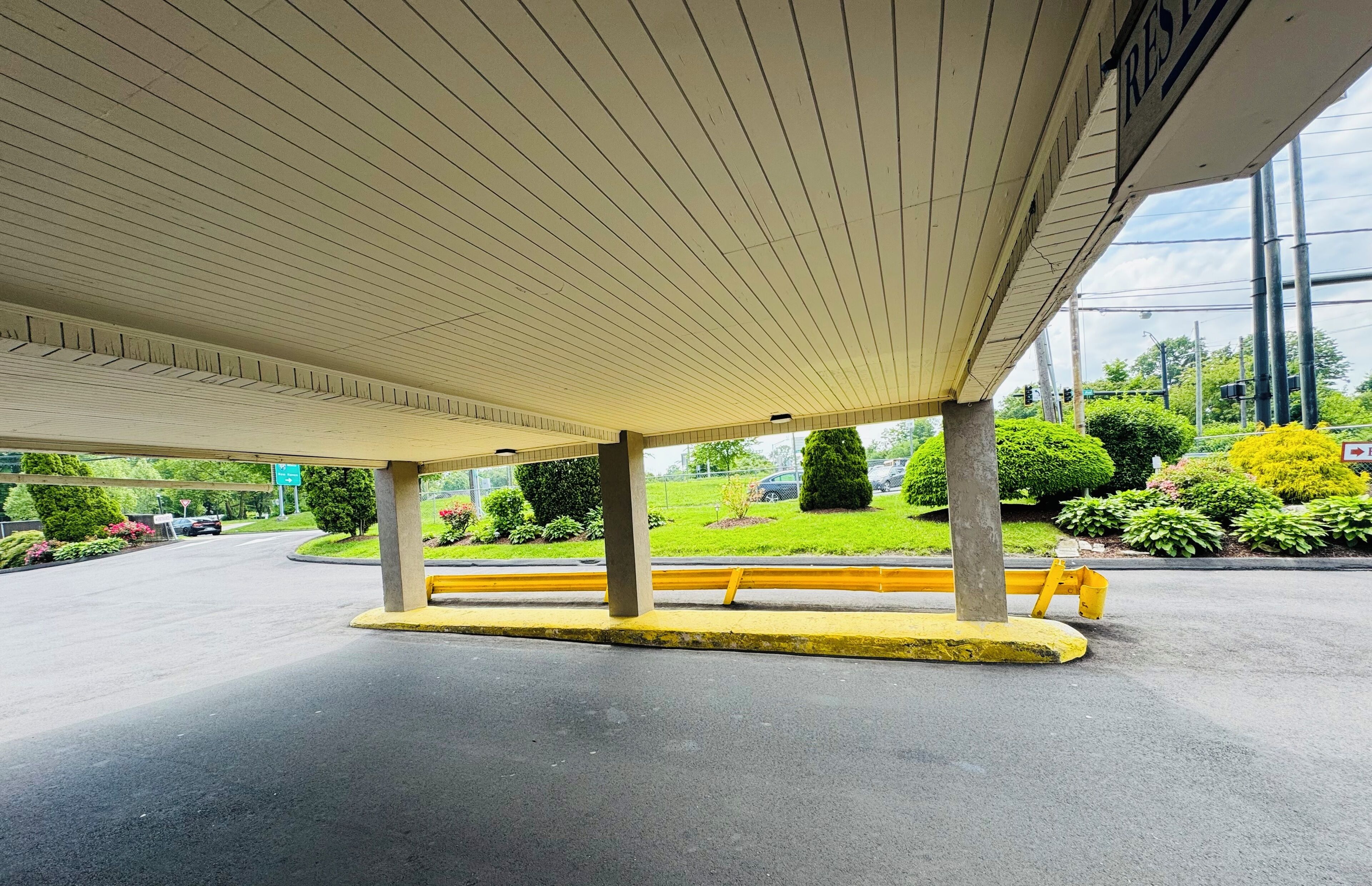 A covered parking area with a bench, concrete pillars, and a view of a landscaped area with greenery and a road.