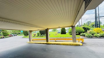 A covered parking area with a bench, concrete pillars, and a view of a landscaped area with greenery and a road.