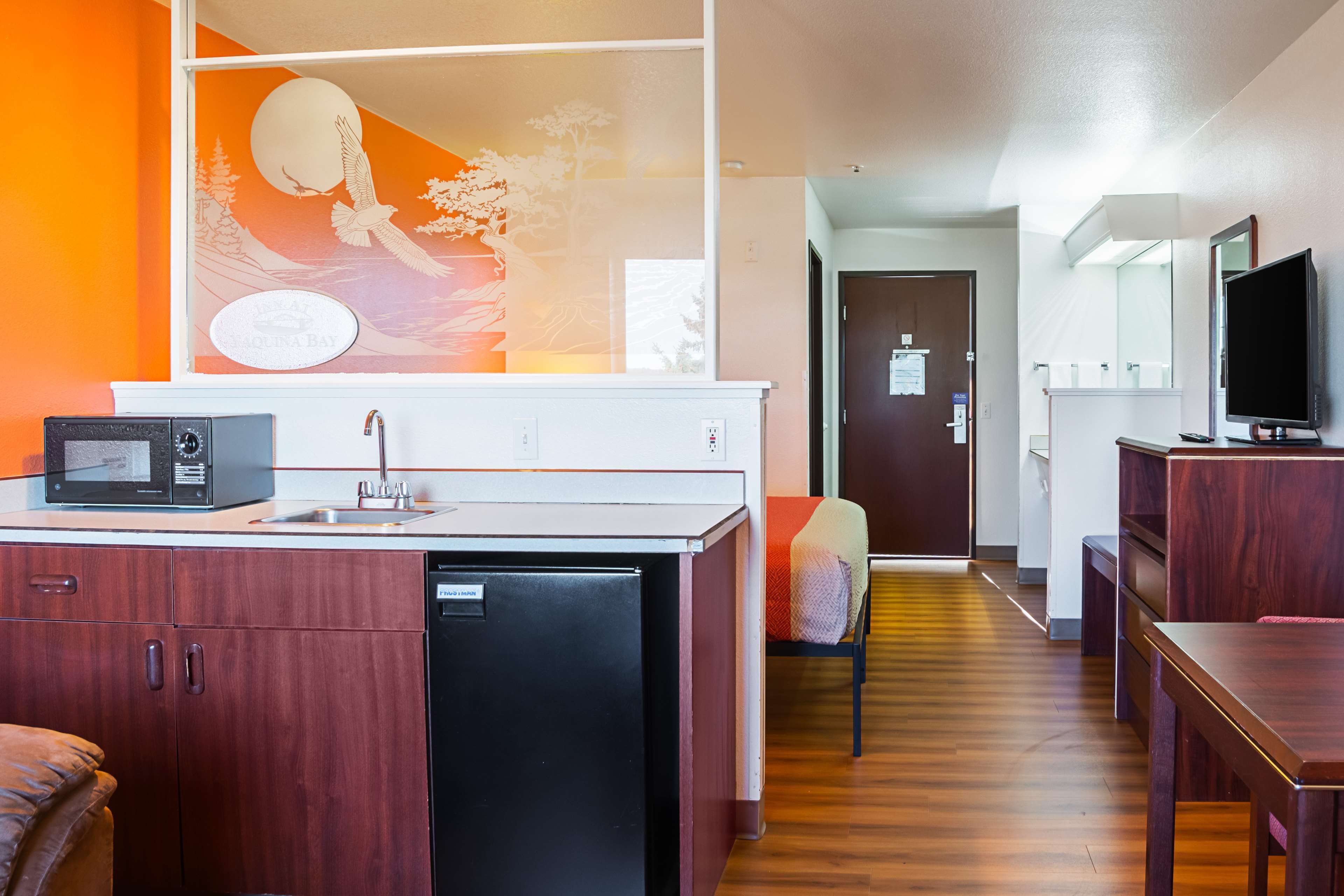 A compact hotel room with a kitchenette, a bed, a television, and a view of a cityscape through the window.