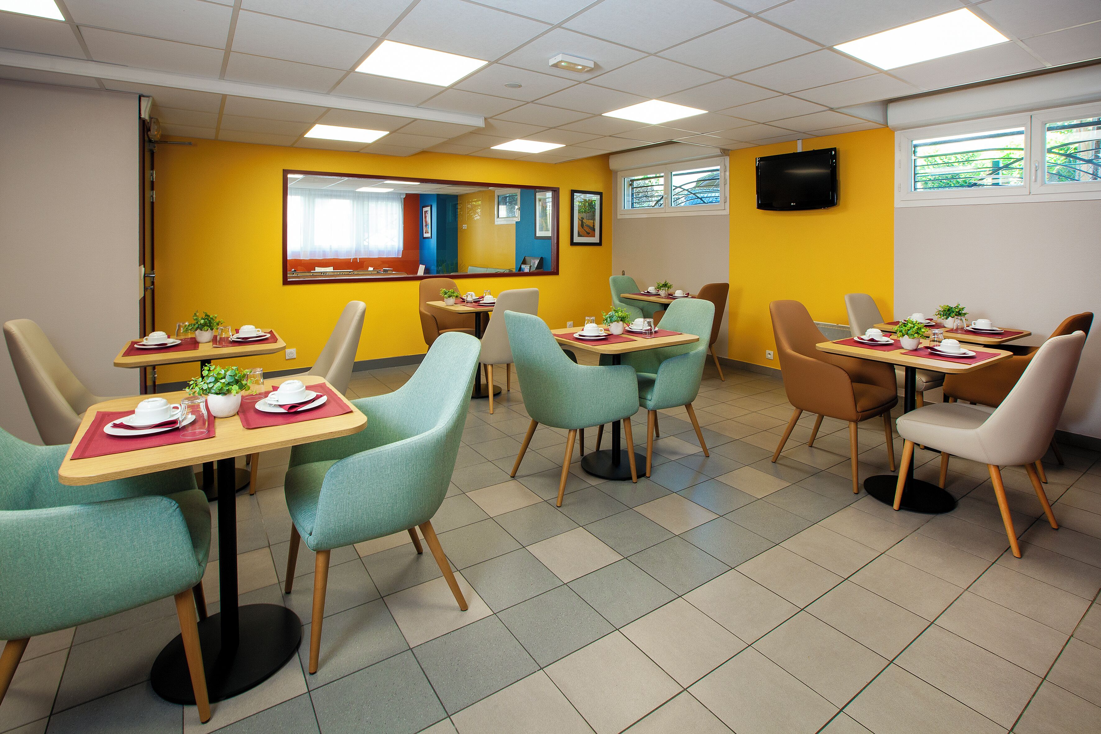 A dining area with tables and chairs, a television mounted on the wall, and a window with a view of the outside.