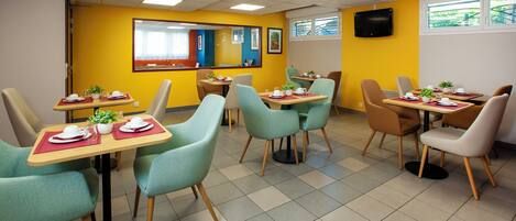 A dining area with tables and chairs, a television mounted on the wall, and a window with a view of the outside.