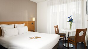 Una habitación de hotel con una cama grande, una mesita de noche con lámpara, una pequeña mesa redonda con sillas y un jarrón con flores.
