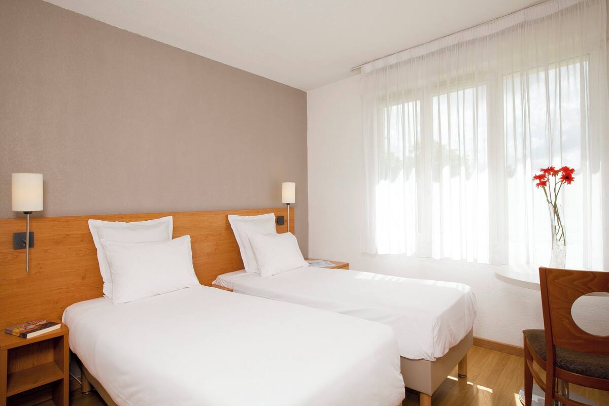 A hotel room with two single beds, wooden headboards, bedside tables with lamps, a wooden chair, and a window with sheer curtains.
