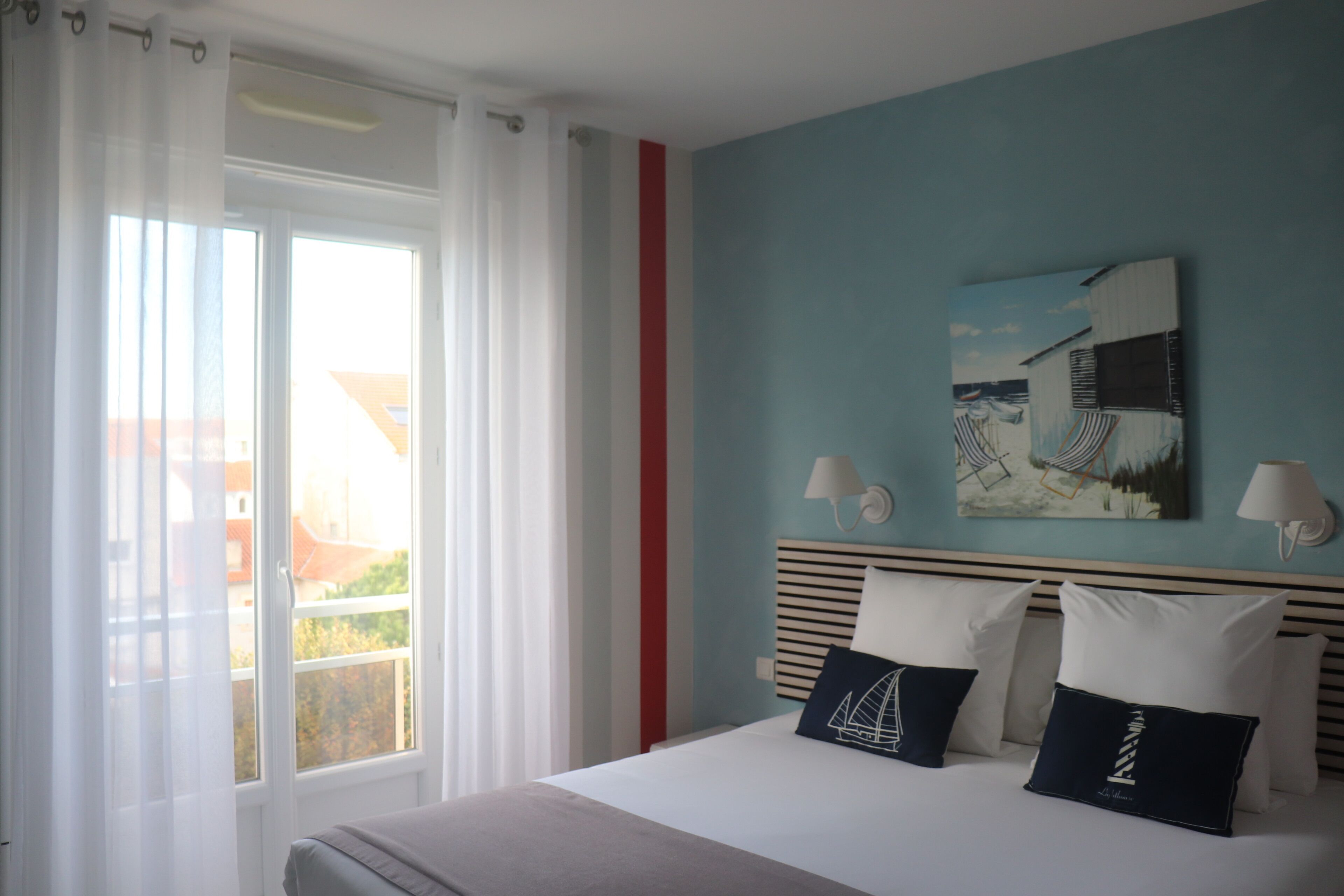 A hotel room with a bed, a painting on the wall, and a view of buildings outside the window.
