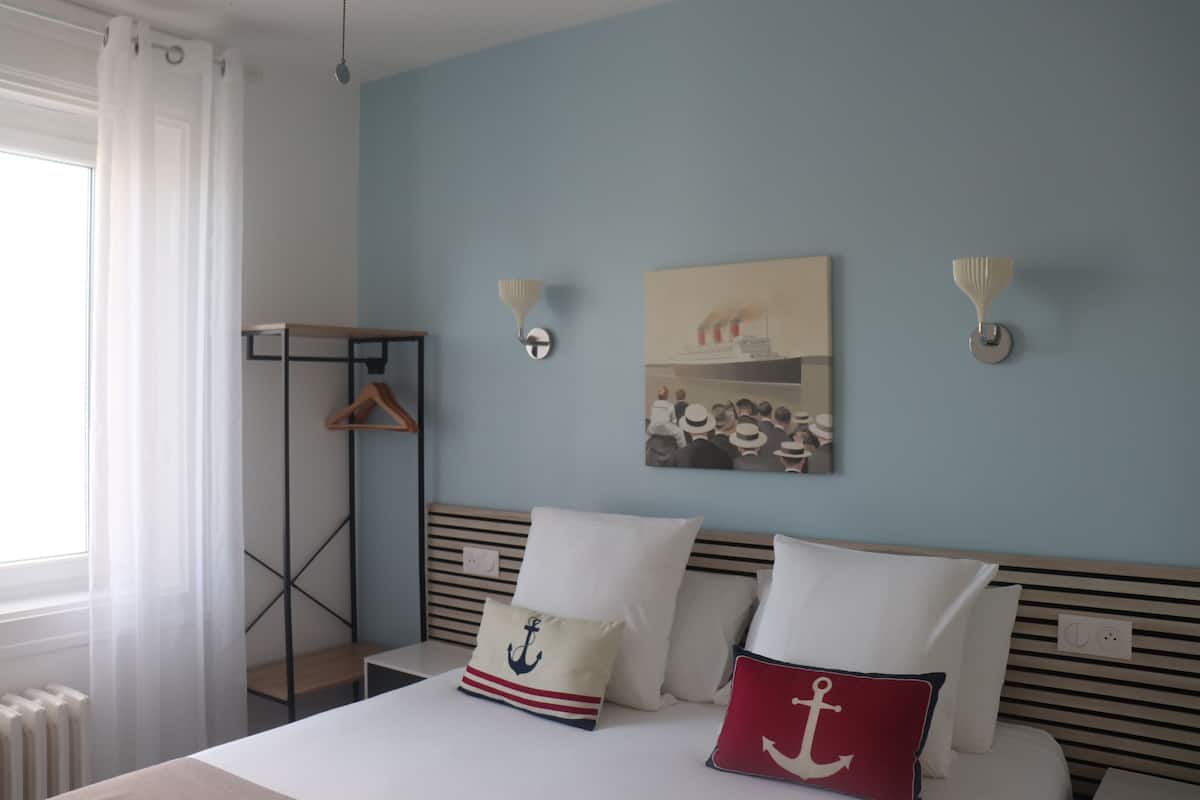 A hotel room with a bed, a hanging coat rack, a wall-mounted light fixture, and a painting.