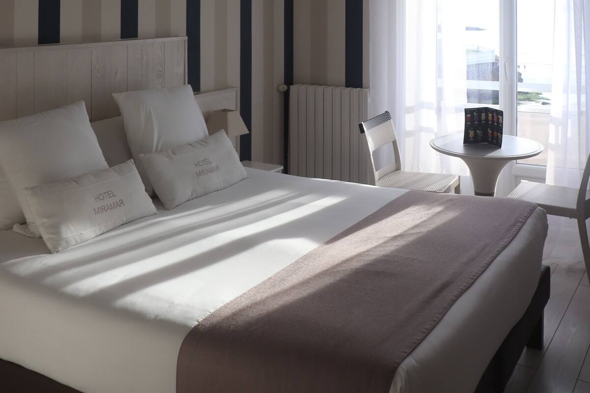 A hotel room with a bed, a ceiling fan, a window with curtains, a chair, and a small table.