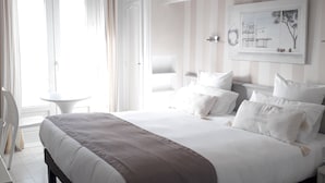 Moderni hotellihuone, jossa on suuri sänky, työpöytä ja tuoli. Huoneessa on ikkuna, jossa on verhot, seinälle kiinnitetty taideteos ja raidallinen seinäkuvio.