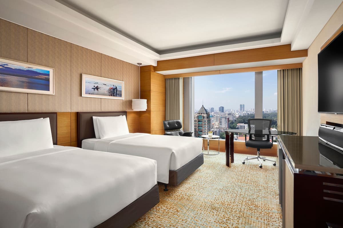 A hotel room with two beds, a desk, a chair, a TV, and a large window with a city view.