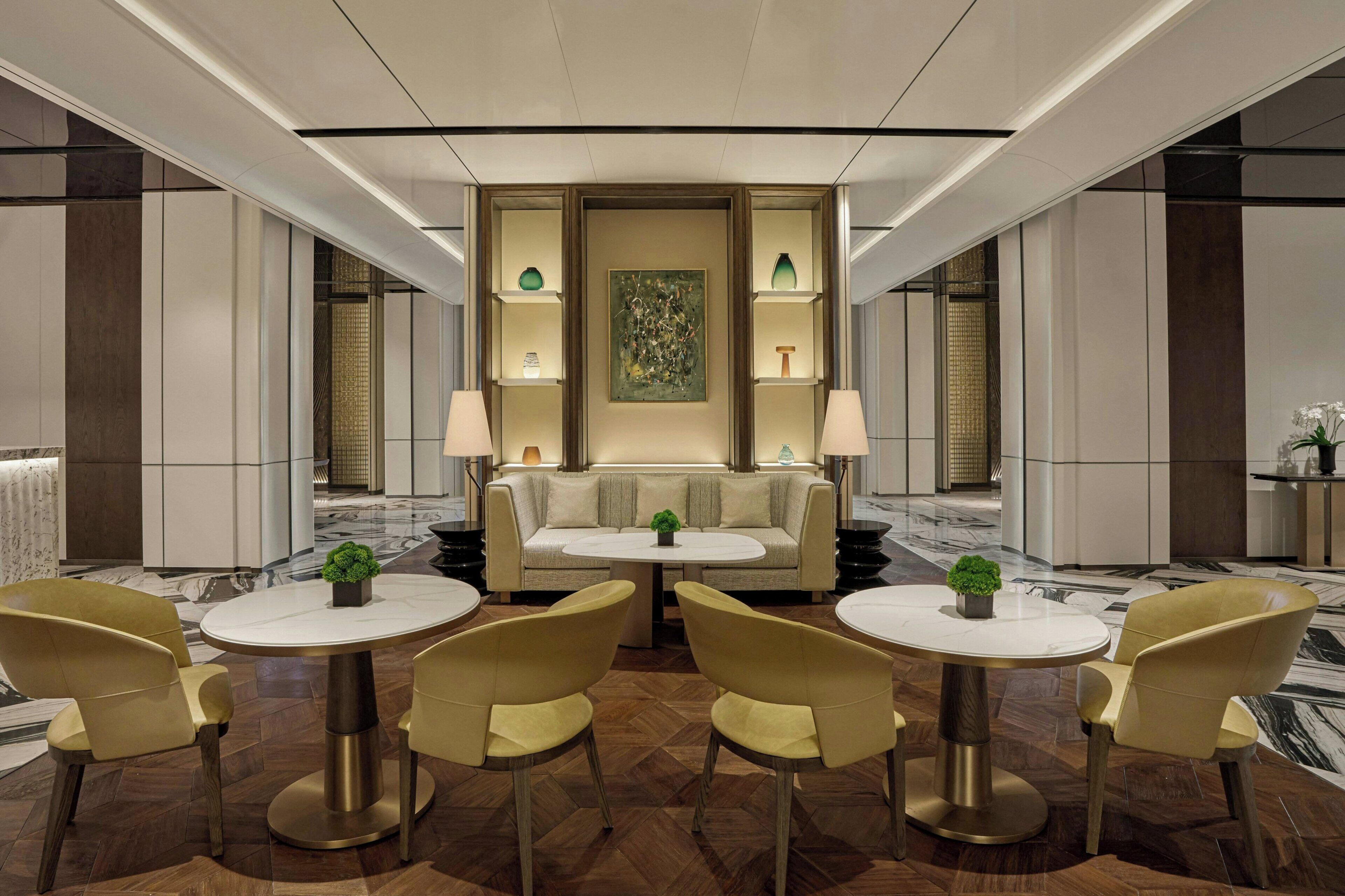 A modern hotel lobby with a central seating area, round tables, and a decorative wall.