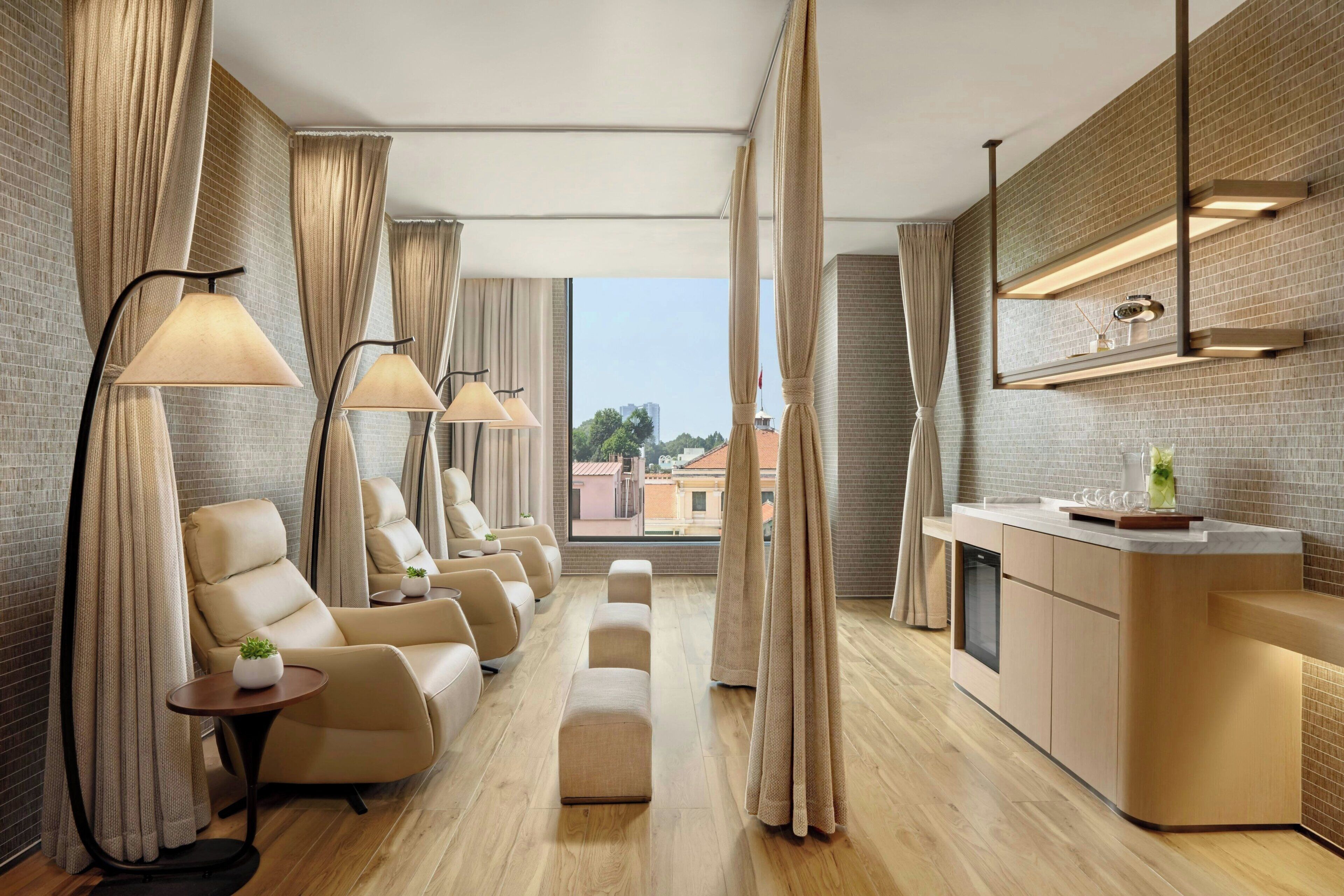 A modern hotel room with a long sofa, a small table, a kitchen area, and large windows.