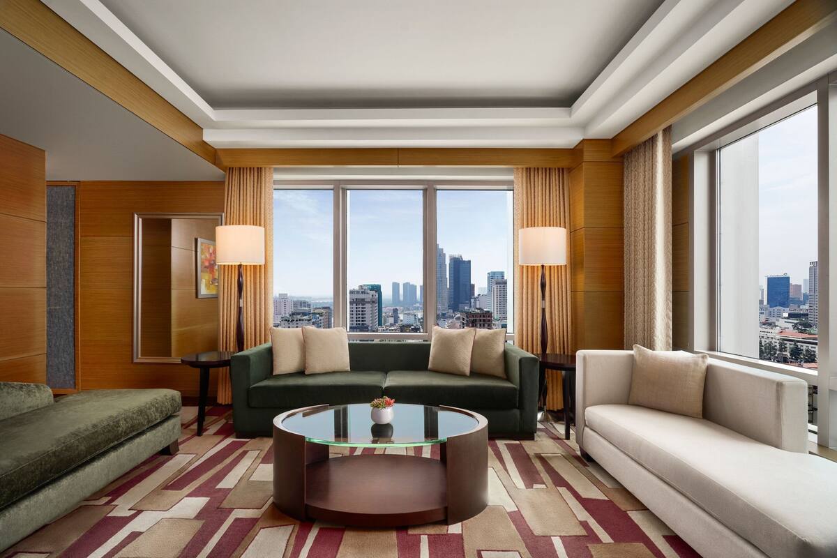 A modern hotel room with a green sofa, a beige sofa, and a glass coffee table. There are large windows with a city view.