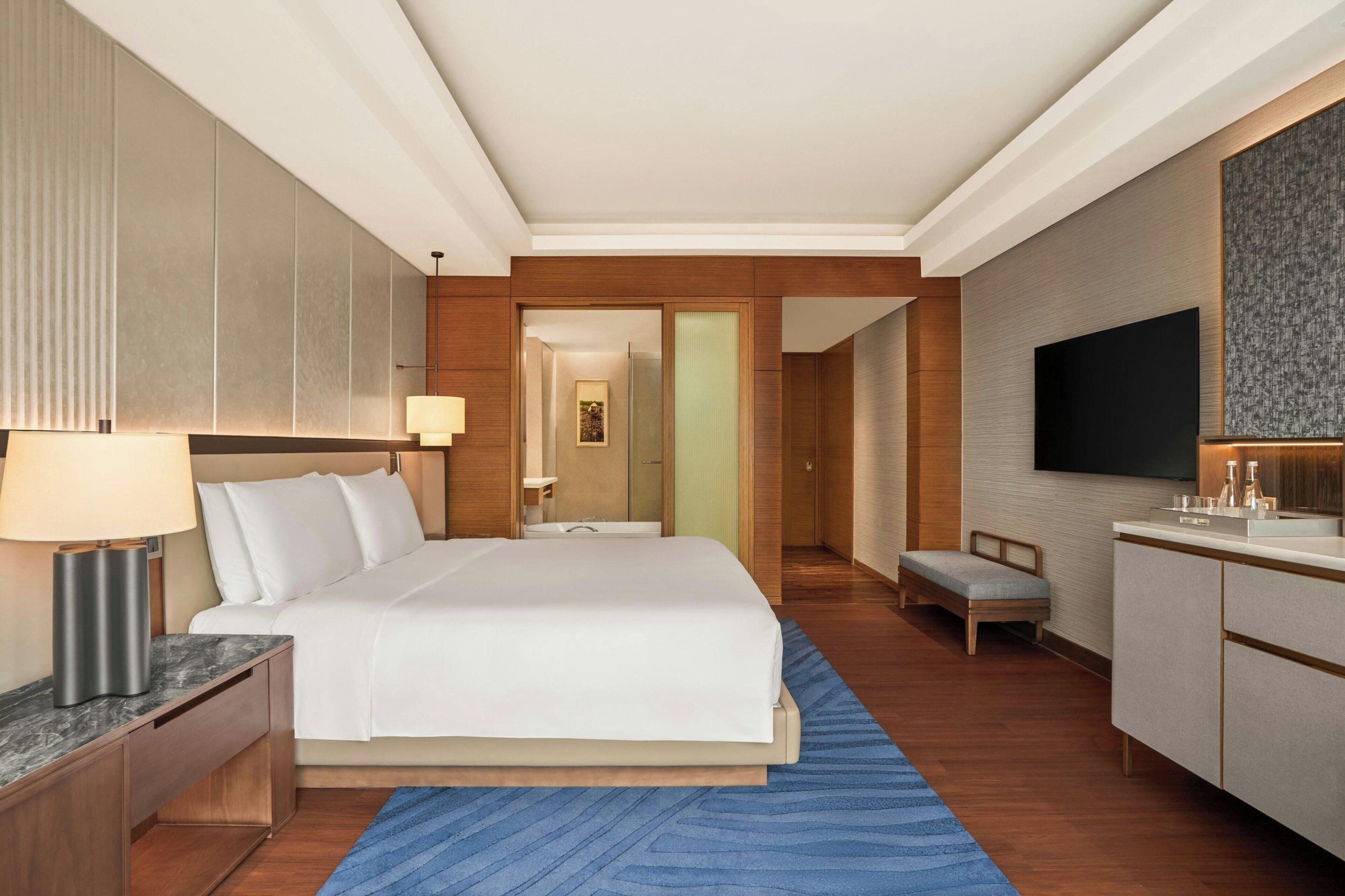 A modern hotel room with a large bed, bedside tables, a desk, a television, and a bathroom visible through an open door.