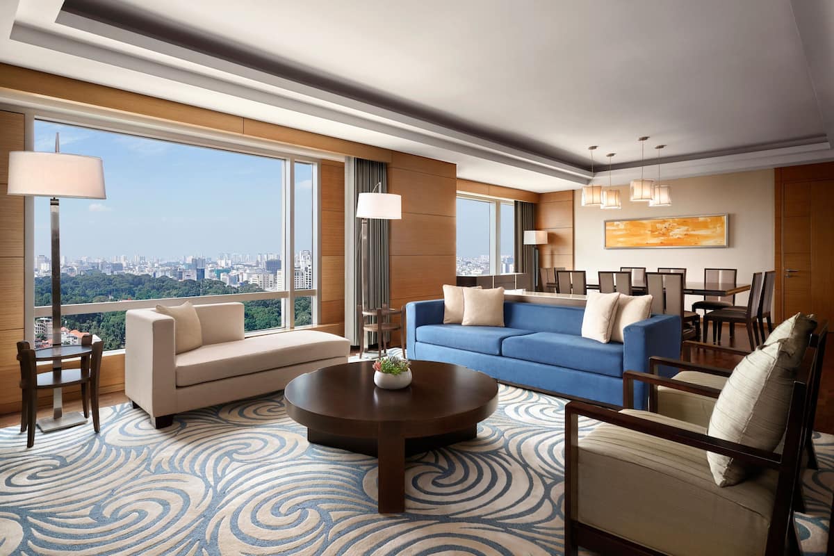 A spacious living room with a large window offering a city view, a blue sofa, a round coffee table, and a patterned rug.