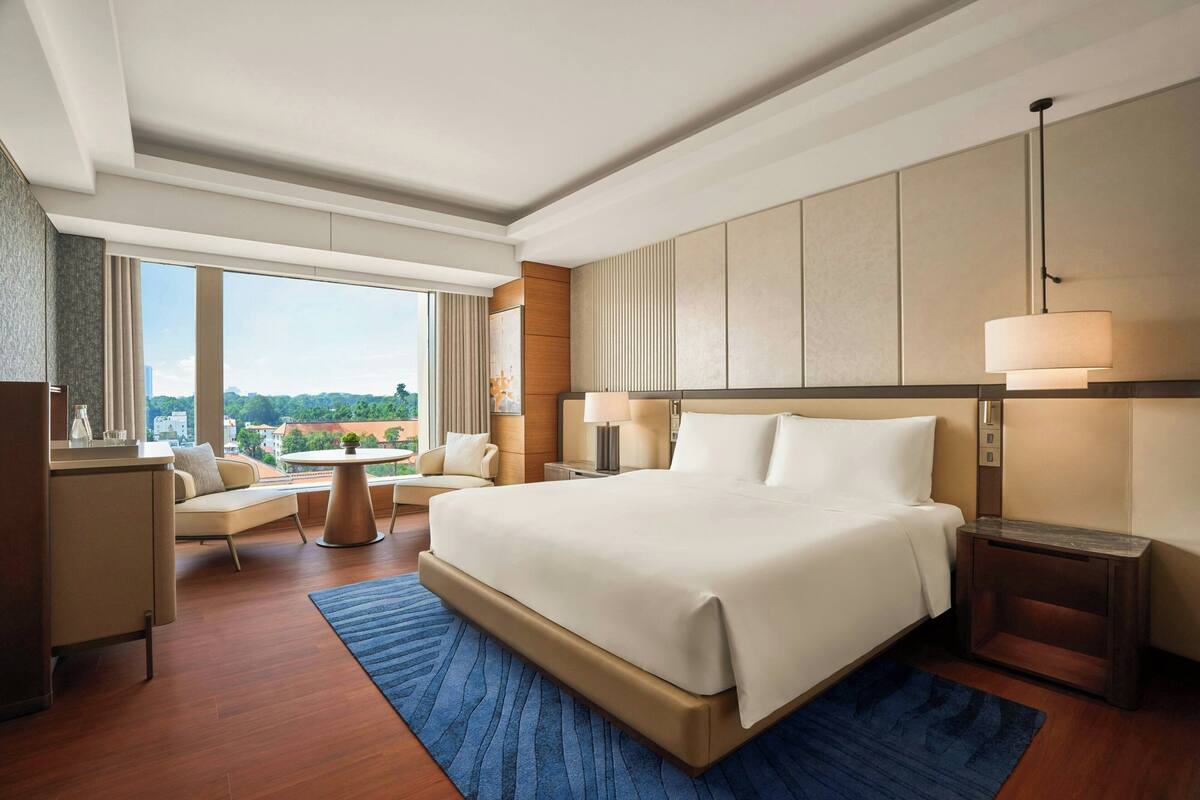 A modern hotel room with a large bed, a desk, a chair, a round table, and a view of the city.