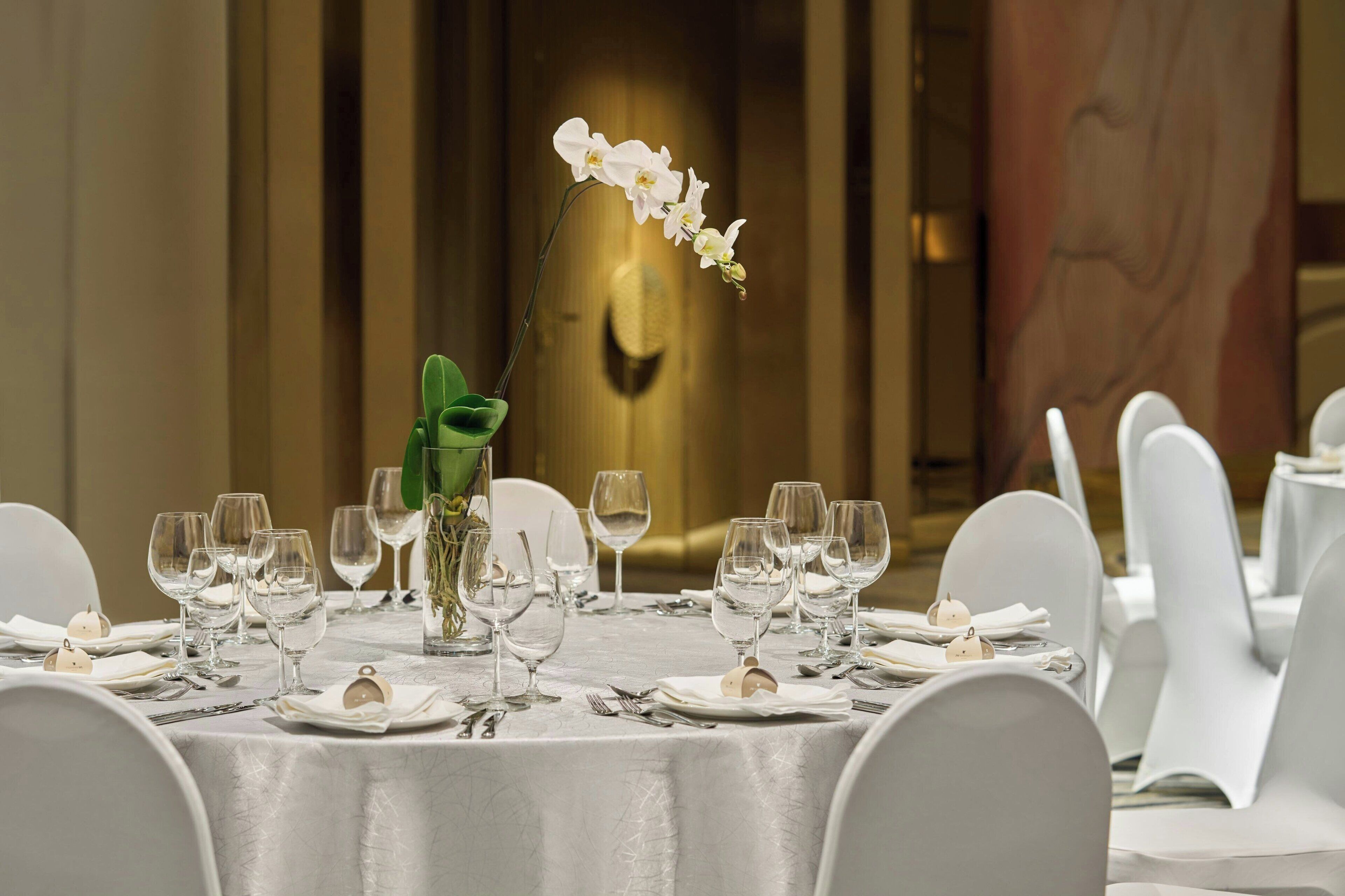 A dining table set with white chairs, wine glasses, and a vase with a white orchid.