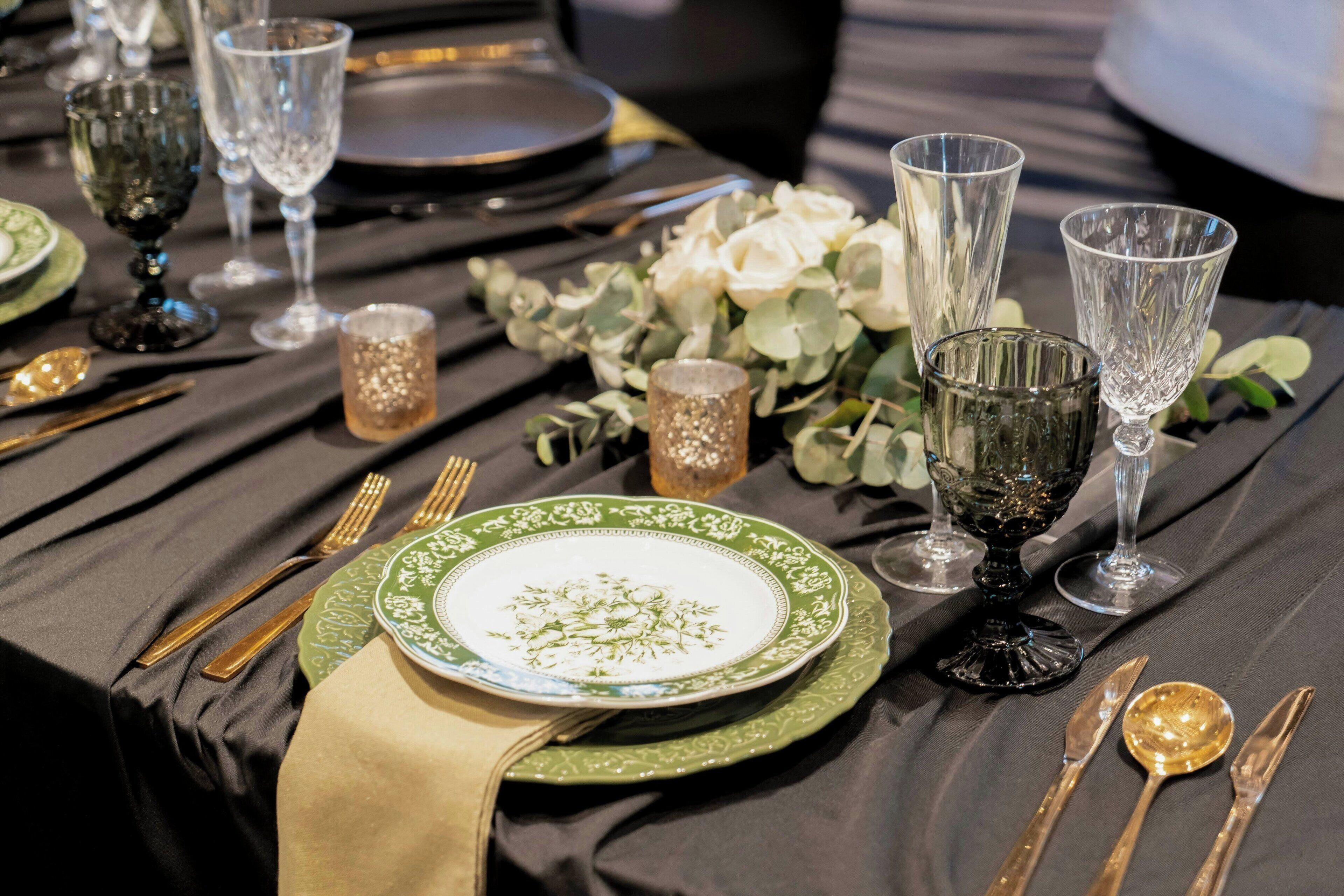 A table set with green and white plates, gold cutlery, and clear glassware, accompanied by small glass bottles and white flowers.