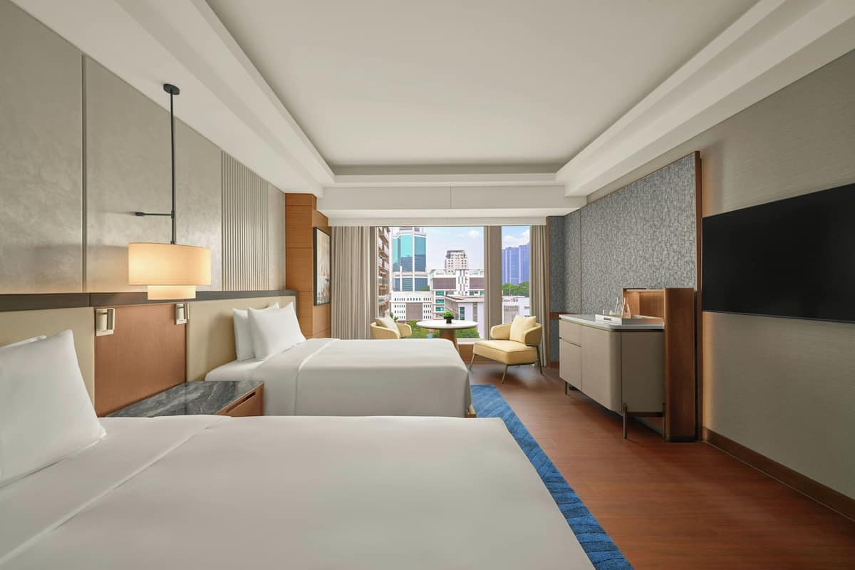 A hotel room with two beds, a desk, a chair, a TV, and a view of the city.