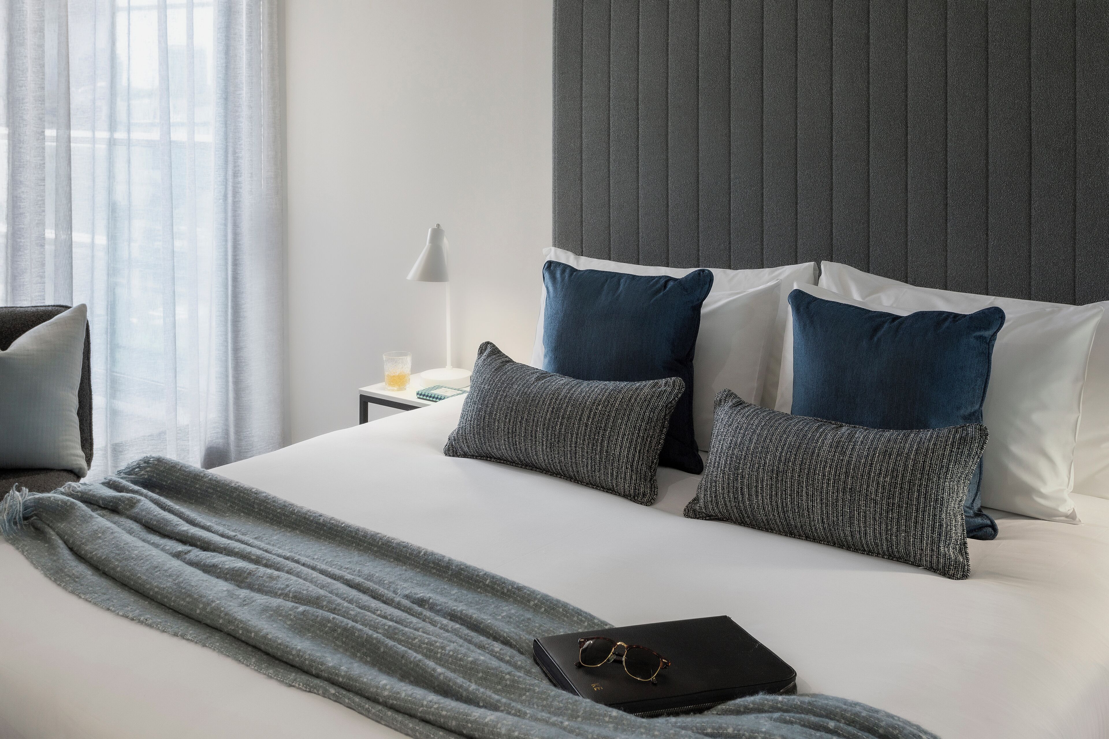 A neatly made bed with a grey blanket, two pillows, and a pair of glasses on a black bag.