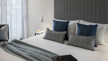 A neatly made bed with a grey blanket, two pillows, and a pair of glasses on a black bag.