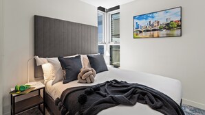 A modern hotel room with a large bed, a nightstand, a lamp, and a cityscape painting on the wall.