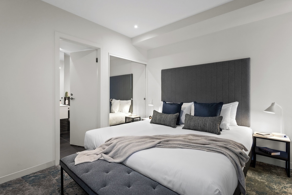 A modern hotel room with a large bed, a bedside table with a lamp, and a mirror above the bed.