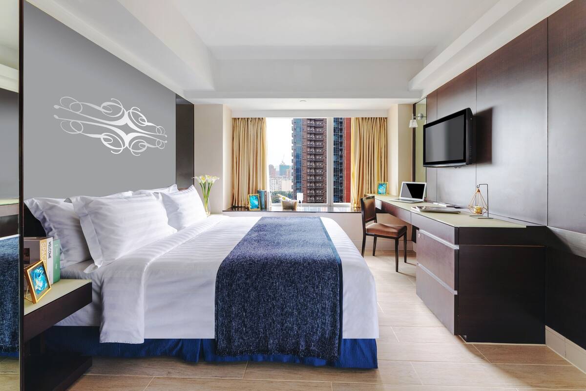 A modern hotel room with a large bed, a desk, a TV, and a view of tall buildings.