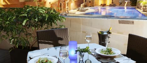 Mesa lista con platos, cubiertos y un vaso de agua, con una piscina y asientos al aire libre al fondo.