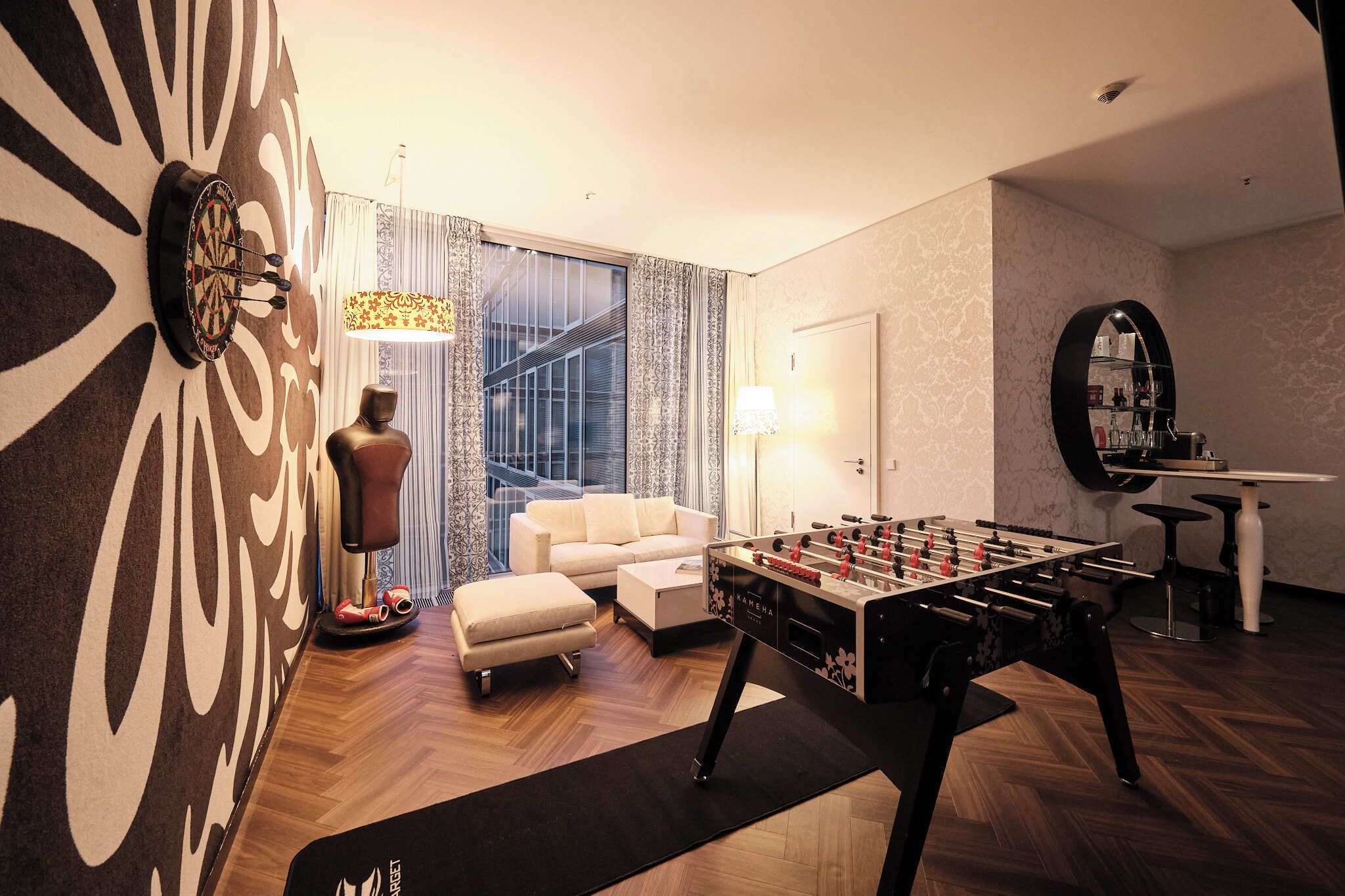 Un salon moderne comprenant une table de baby-foot, un canapé, une table basse, un jeu de fléchettes et une grande décoration murale abstraite.