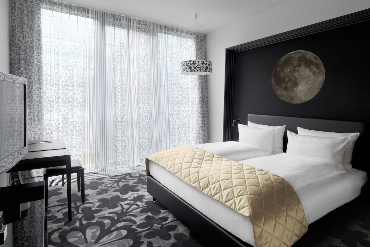 A modern hotel room with a large bed, a desk, and a large window with sheer curtains.