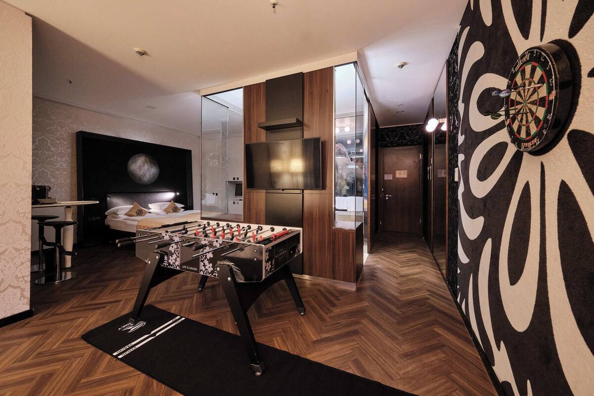 A modern hotel room with a foosball table, a dartboard, a TV, and a large mirror.