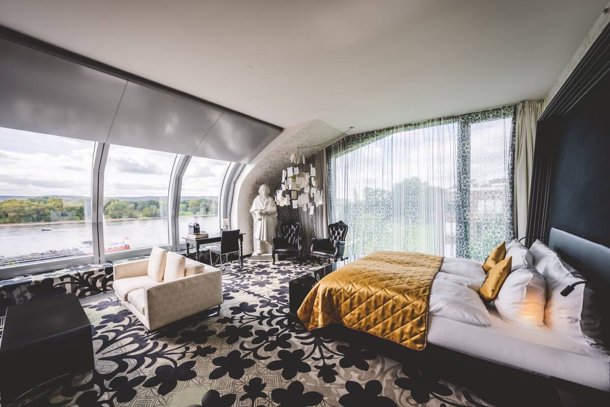 A modern hotel room with a large bed, a sofa, a desk, and a view of a river.